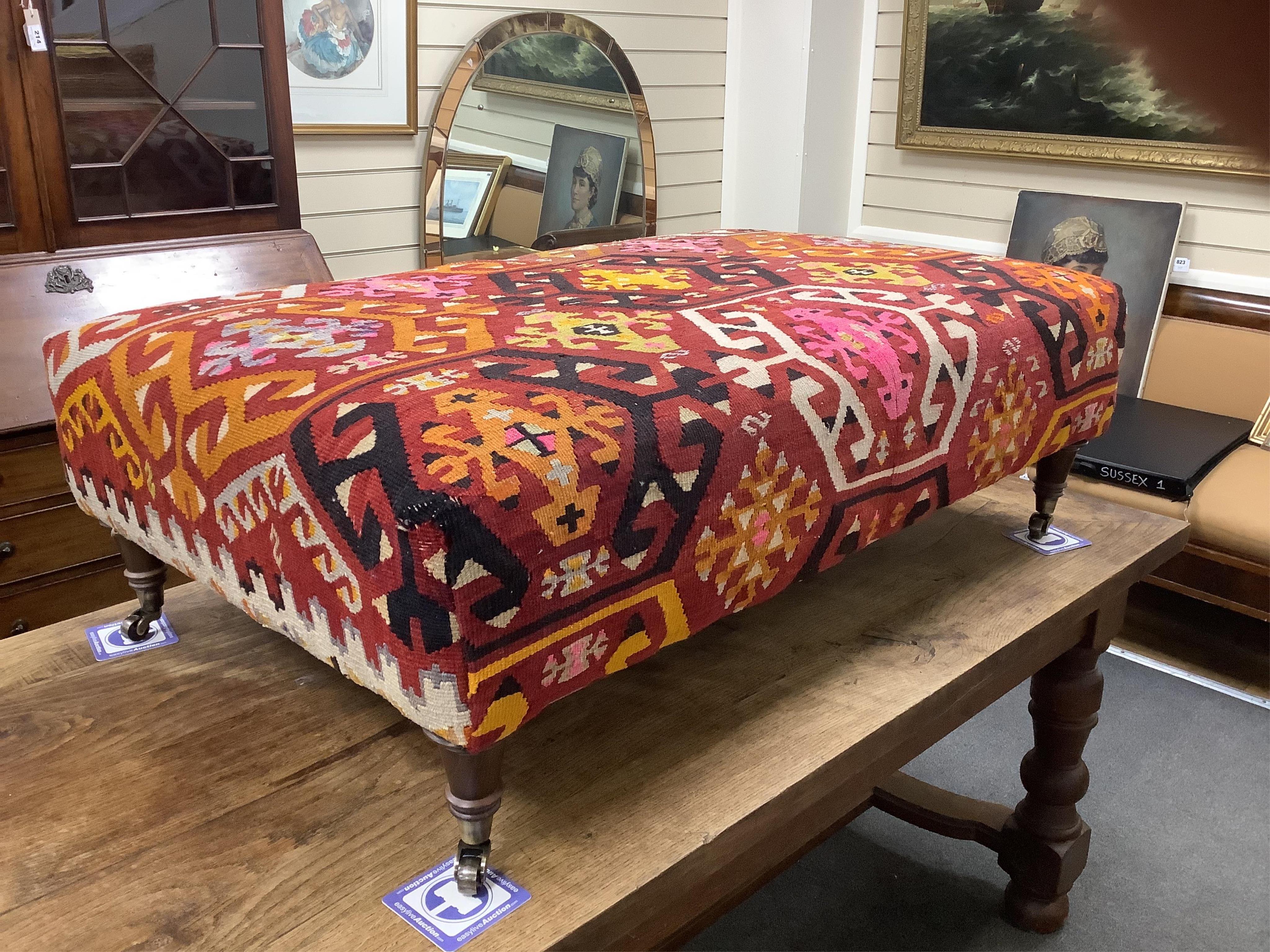 A large contemporary rectangular footstool with antique polychrome Kilim upholstery on turned beech feet with brass caps and castors, width 124cm, depth 78cm, height 40cm.